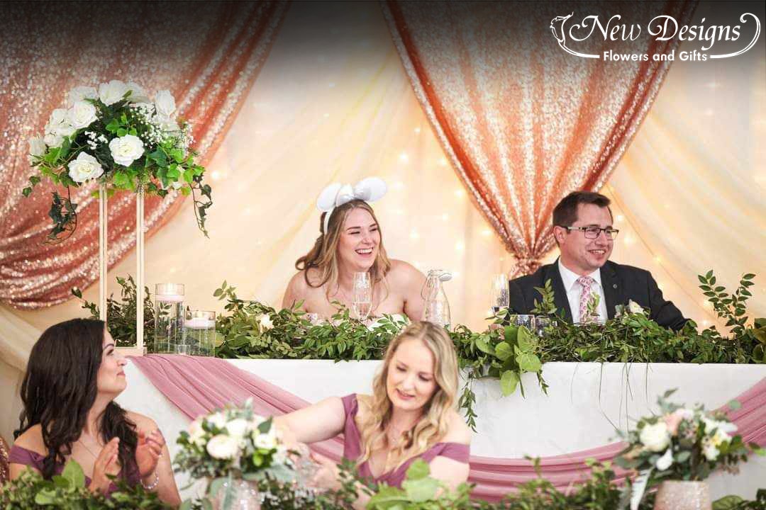 A bride and grooms it at their 2 tiered head table, lined with Greenery and blush and cream roses in front of A Cream and Rose gold Backdrop